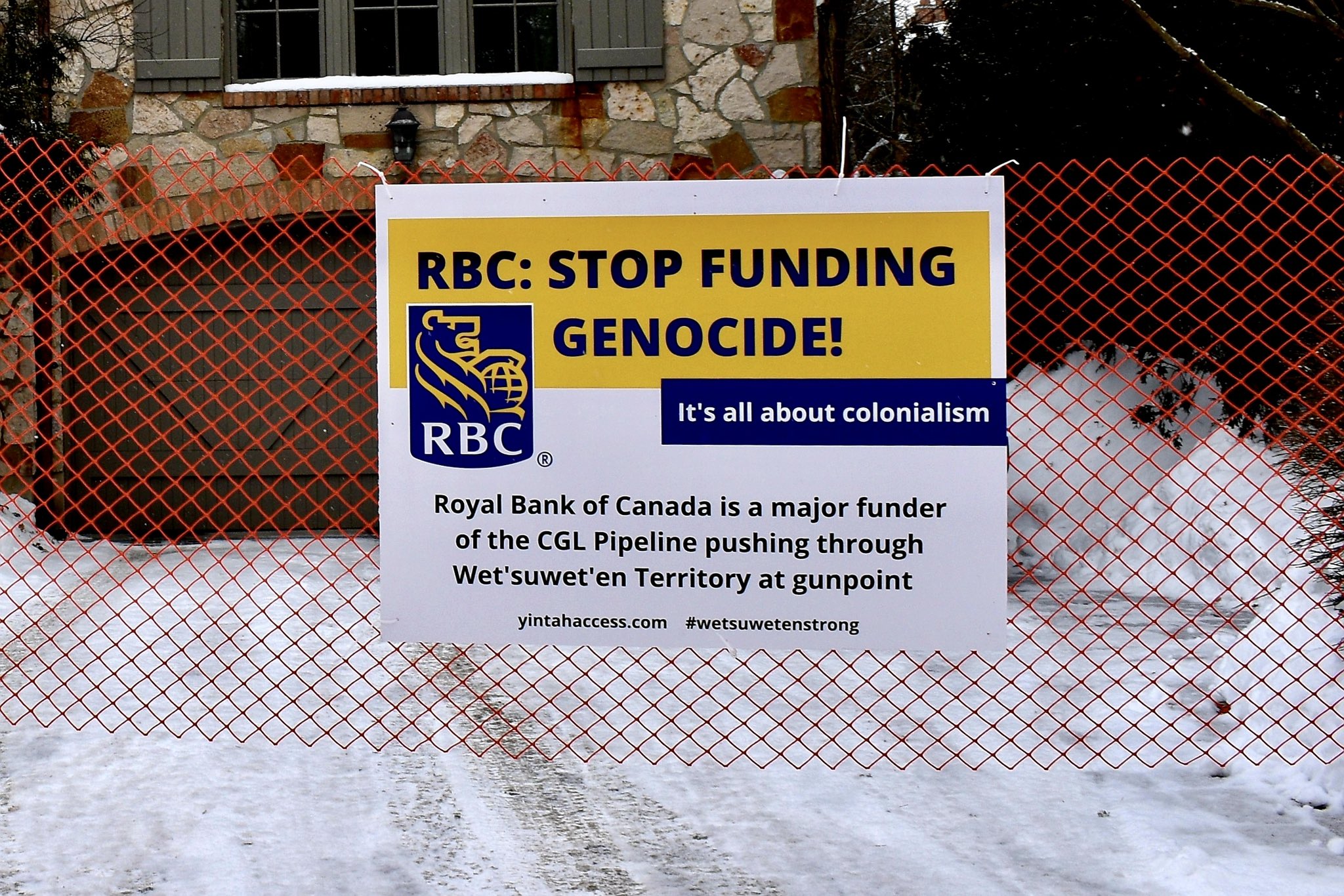 A sign is placed on orange construction mesh in front Doug Guzman's house, reading "RBC: STOP FUNDING GENOCIDE!" Photo credit: World BEYOND War Canada