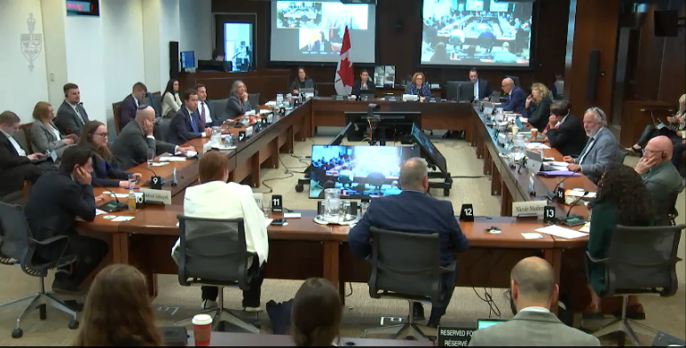 Committee in session at the House of Commons on May 9, 2024. Over a dozen people are sitting facing each other at tables in a rectangular shape.