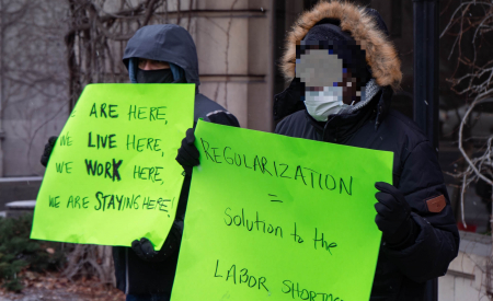 Two racialized people hold up signs outside in winter. One is light green with black text reading "Regularisation = solution to the labour shortage." Photo from a Solidarity Across Borders event in December 2021 in Montreal, Undocumented migrants have a solution to the labor shortage: Status for all. Photo: Solidarity Across Borders.